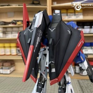 May include: A detailed model robot kit with black, red, white, and blue detailing. The model features intricate paneling and design elements, with the text "AG" visible on the side. The model is set against a blurred background.