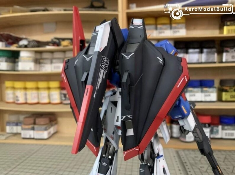 May include: A detailed model robot kit with black, red, white, and blue detailing. The model features intricate paneling and design elements, with the text "AG" visible on the side. The model is set against a blurred background.