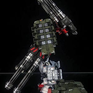 K&ouml;nnte beinhalten: Ein detaillierter Roboter-Modellbausatz in den Farben Wei&szlig;, Rot und Olivgr&uuml;n. Die Figur ist mit zwei gro&szlig;en Gatling-Gesch&uuml;tzen mit mehreren L&auml;ufen und mehreren Raketenwerfern ausgestattet. Der Text AeroModelBuild ist in der Ecke zu sehen.