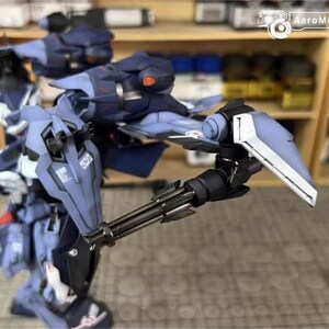 May include: A detailed, partially assembled model kit of a futuristic robot, primarily in shades of blue and gray. The robot features intricate mechanical details, including a multi-barreled gun and angular armor plating. The model is set against a backdrop of shelves with paint bottles.