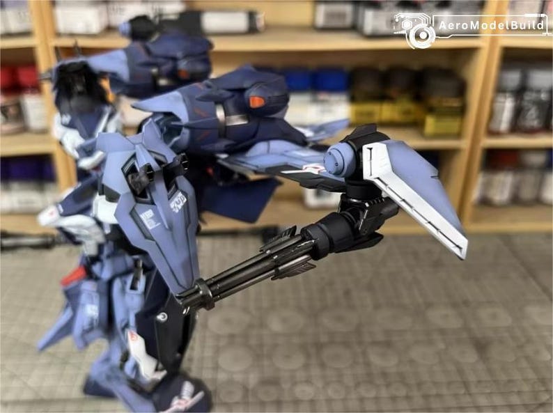 May include: A detailed, partially assembled model kit of a futuristic robot, primarily in shades of blue and gray. The robot features intricate mechanical details, including a multi-barreled gun and angular armor plating. The model is set against a backdrop of shelves with paint bottles.