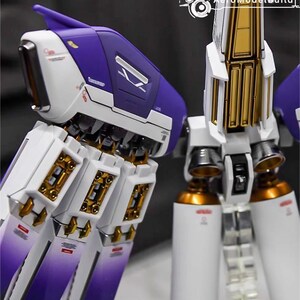 May include: Close-up of a detailed model with white, purple, and gold accents. The model features intricate mechanical components, including cylinders and panels. The design incorporates sharp angles and metallic finishes, suggesting a futuristic or sci-fi theme.