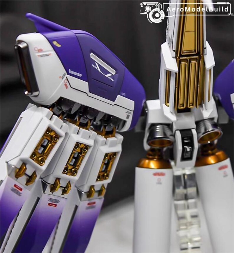 May include: Close-up of a detailed model with white, purple, and gold accents. The model features intricate mechanical components, including cylinders and panels. The design incorporates sharp angles and metallic finishes, suggesting a futuristic or sci-fi theme.