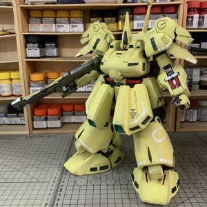 May include: A large, assembled model of a yellow and green mech robot with a long gun. The model has intricate details, including panel lines, decals, and articulated joints. The model is set against a backdrop of shelves with paint supplies.