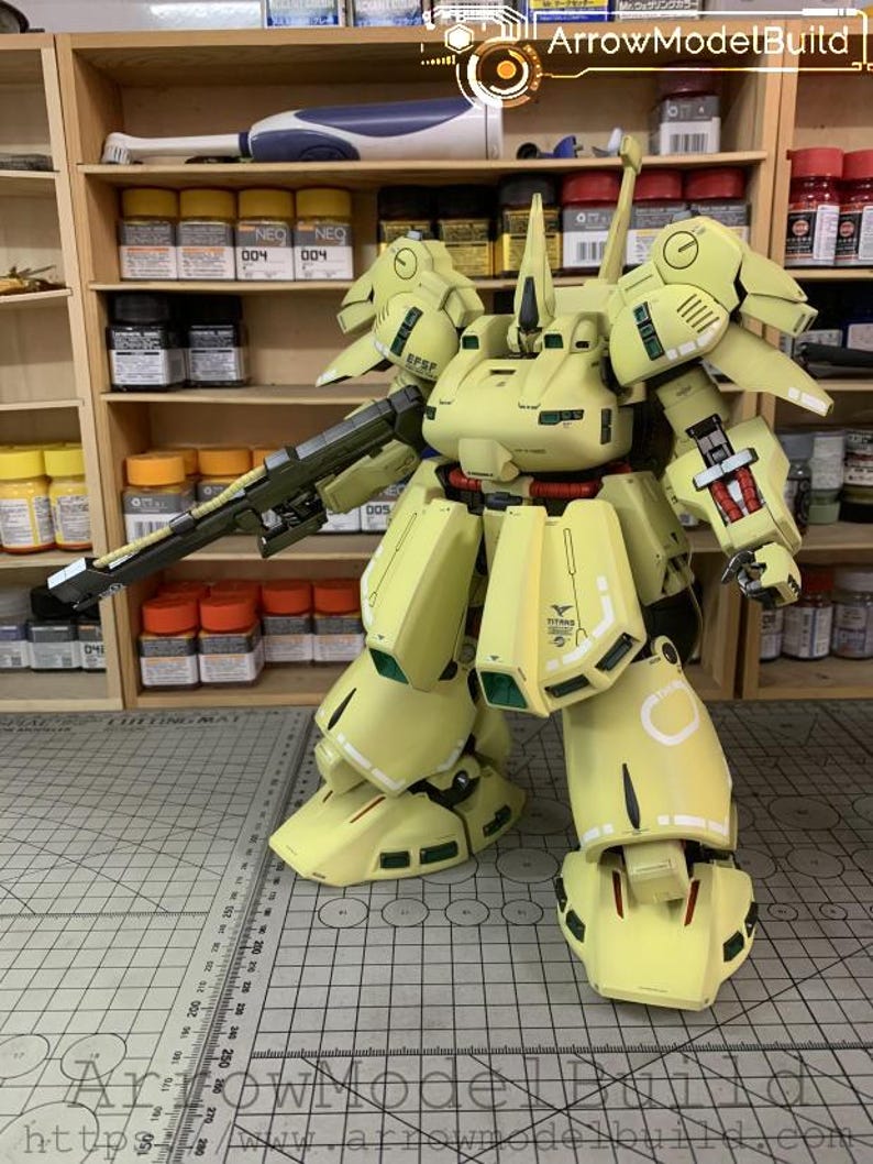 May include: A large, assembled model of a yellow and green mech robot with a long gun. The model has intricate details, including panel lines, decals, and articulated joints. The model is set against a backdrop of shelves with paint supplies.