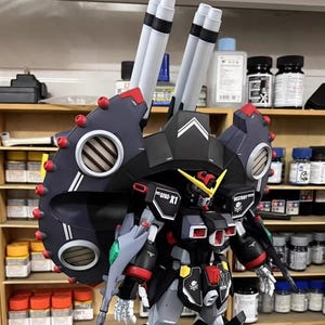 May include: A detailed model of a black, red, and gray mecha robot with large shoulder-mounted cannons. The robot has intricate detailing, including vents, panels, and the text "GFAS-X1" and "DESTROY". The model is set against a backdrop of shelves with various containers.
