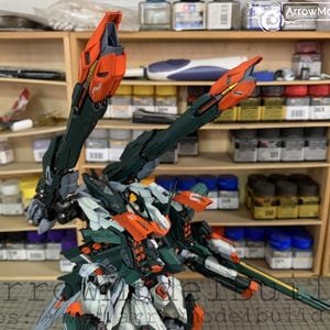 May include: A detailed model kit of a robot with orange, green, white, and black accents. The robot has large, articulated arms and a complex design. The background shows shelves with various model-building supplies.
