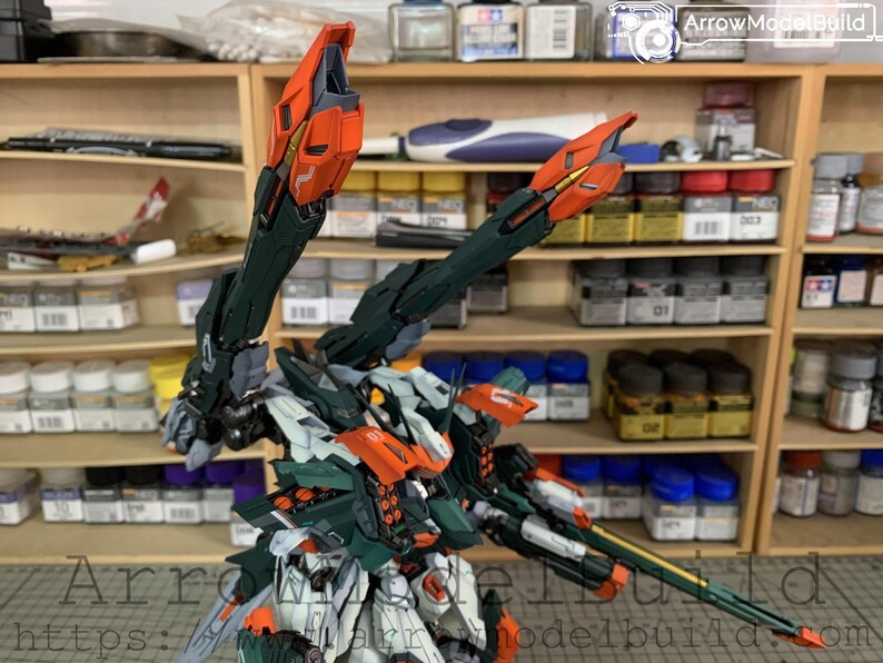 May include: A detailed model kit of a robot with orange, green, white, and black accents. The robot has large, articulated arms and a complex design. The background shows shelves with various model-building supplies.