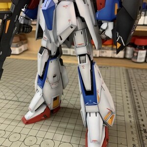 May include: A close-up of a robot model, predominantly white with blue, red, and black detailing. The image highlights the legs, revealing complex design elements. The model is positioned on a grid, with the AeroModelBuild logo visible.