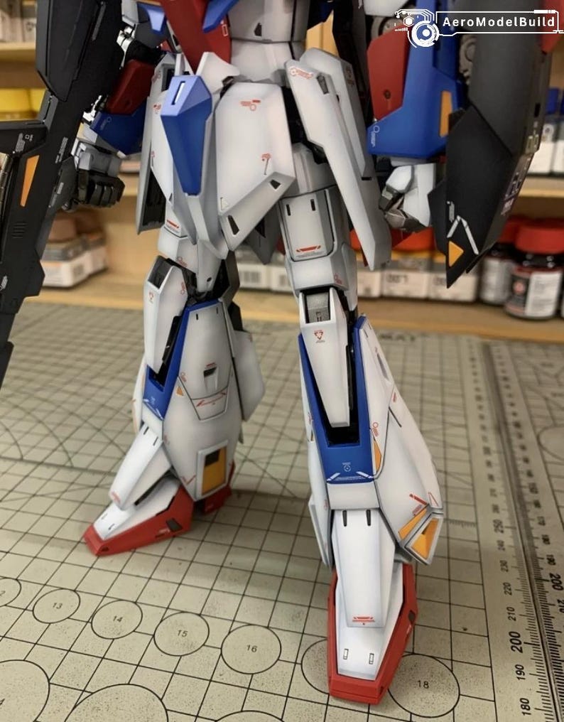 May include: A close-up of a robot model, predominantly white with blue, red, and black detailing. The image highlights the legs, revealing complex design elements. The model is positioned on a grid, with the AeroModelBuild logo visible.