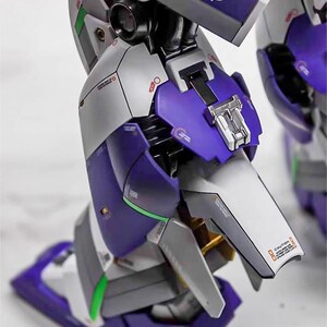 May include: Close-up of a detailed model robot leg. The model features a combination of purple, silver, and white panels with intricate mechanical details. The model has green and gold accents, and the text "CAUTION" is visible.