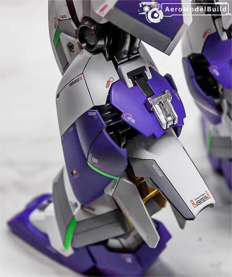 May include: Close-up of a detailed model robot leg. The model features a combination of purple, silver, and white panels with intricate mechanical details. The model has green and gold accents, and the text "CAUTION" is visible.