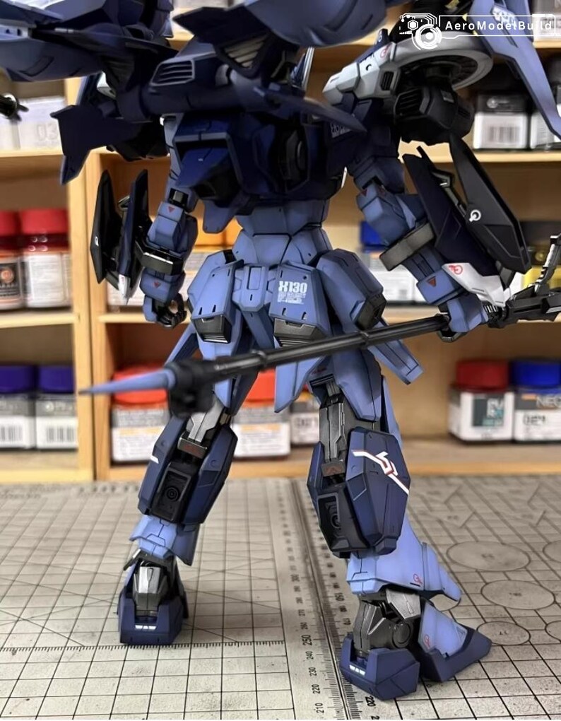 May include: A detailed, assembled model of a robotic figure in shades of blue and black. The model features intricate mechanical details, including wings, weapons, and the text "H130". The figure is posed on a surface with a ruler.