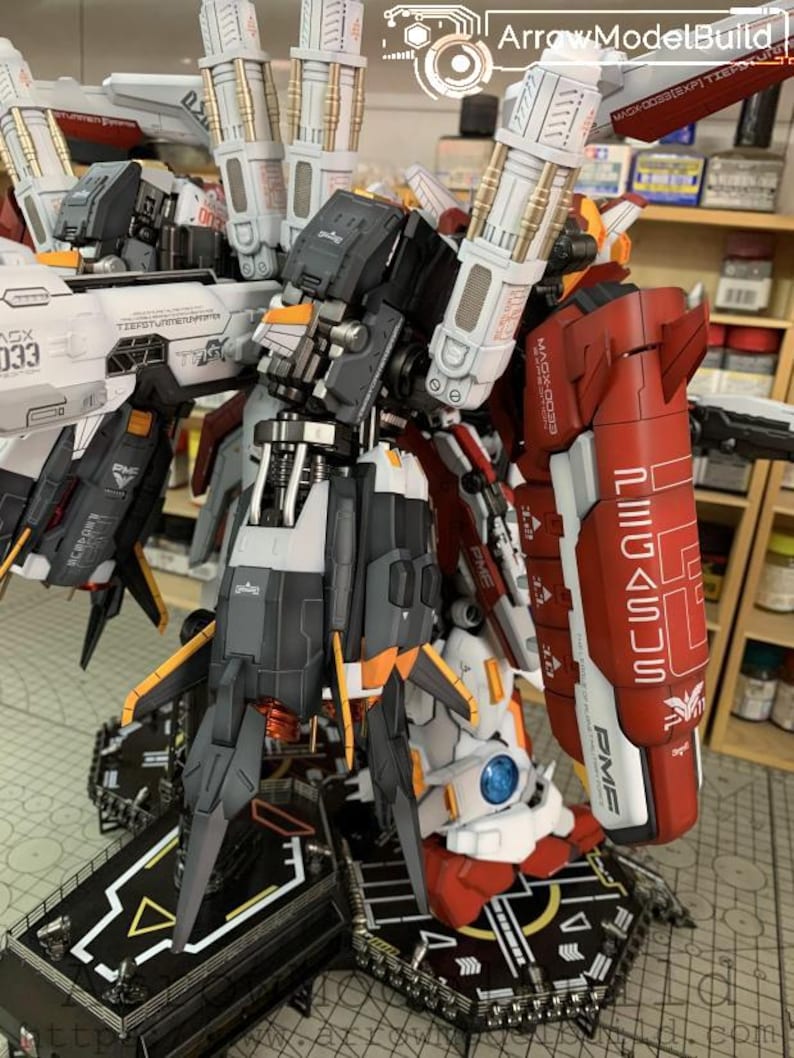 Puede incluir: Un detallado kit de modelo de robot mec&aacute;nico, con una combinaci&oacute;n de colores rojo, blanco, gris y naranja. El modelo tiene intrincados detalles mec&aacute;nicos, incluyendo m&uacute;ltiples paneles, articulaciones y armas. La base es una plataforma negra con marcas blancas y amarillas. El texto "MAGX-0033" es visible.