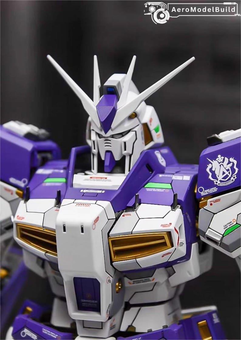 May include: A detailed model of a Gundam robot, featuring a color scheme of white, purple, and gold accents. The robot has a complex design with sharp angles and intricate paneling. The head has two prominent white antennae. The AeroModelBuild logo is in the top right corner.