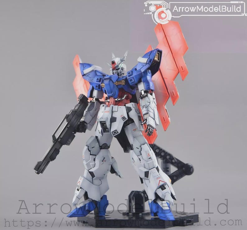 May include: A detailed robot model with a gun, in blue, white, red, and black. The model has large wings and stands on a black base. The text "ArrowModelBuild" is visible.