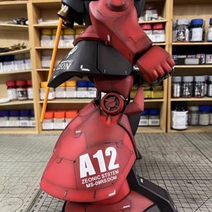 May include: A detailed model of a red and black robot with the text "A12 ZEONIC SYSTEM MS-09RS DOM". The robot has articulated joints and a weathered finish, suggesting a battle-worn appearance. A yellow pole extends from the robot's shoulder.