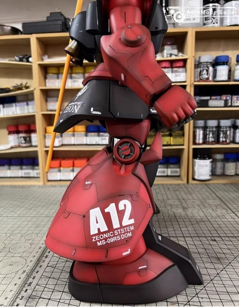 May include: A detailed model of a red and black robot with the text "A12 ZEONIC SYSTEM MS-09RS DOM". The robot has articulated joints and a weathered finish, suggesting a battle-worn appearance. A yellow pole extends from the robot's shoulder.