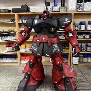 May include: A detailed model of a red and black robot with the text "ZEON" on the skirt armor. The robot has a distinctive head design and is posed with arms extended. The model is set against a backdrop of shelves with various bottles and containers.