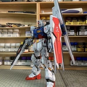 K&ouml;nnte beinhalten: Detailliertes Modell eines Gundam-Roboters, haupts&auml;chlich wei&szlig; mit roten, blauen und gelben Akzenten. Es verf&uuml;gt &uuml;ber einen gro&szlig;en Schild mit dem Text "EFSF" und ein langes Gewehr. Das Modell steht in einer Werkstatt mit Regalen voller Farben und Werkzeuge.