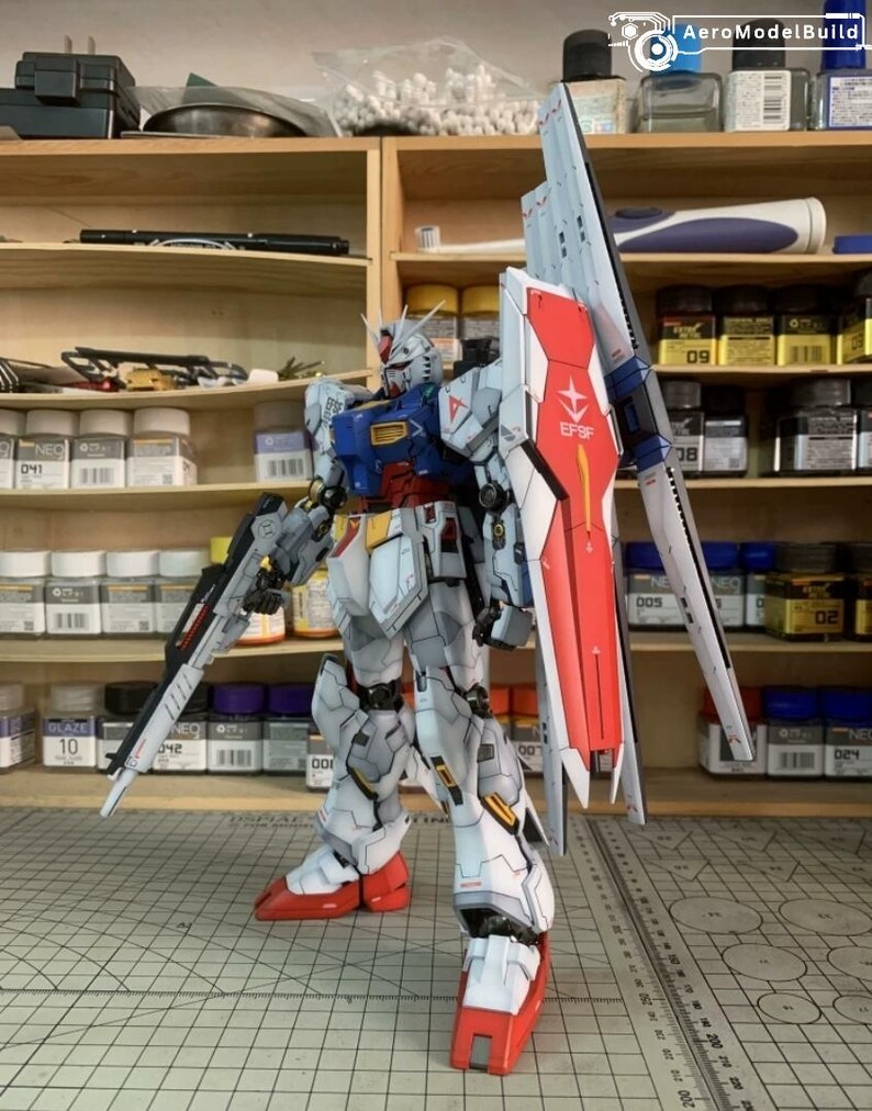 K&ouml;nnte beinhalten: Detailliertes Modell eines Gundam-Roboters, haupts&auml;chlich wei&szlig; mit roten, blauen und gelben Akzenten. Es verf&uuml;gt &uuml;ber einen gro&szlig;en Schild mit dem Text "EFSF" und ein langes Gewehr. Das Modell steht in einer Werkstatt mit Regalen voller Farben und Werkzeuge.