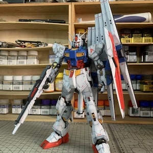 K&ouml;nnte beinhalten: Ein detailliertes Modell eines Gundam-Roboters, haupts&auml;chlich in Wei&szlig;, Blau, Rot und Gelb. Der Roboter ist mit einer gro&szlig;en Waffe und Fl&uuml;geln posiert und weist komplizierte Linien und Abziehbilder auf. Das Modell steht vor einem Hintergrund von Regalen mit verschiedenen Hobbyartikeln.