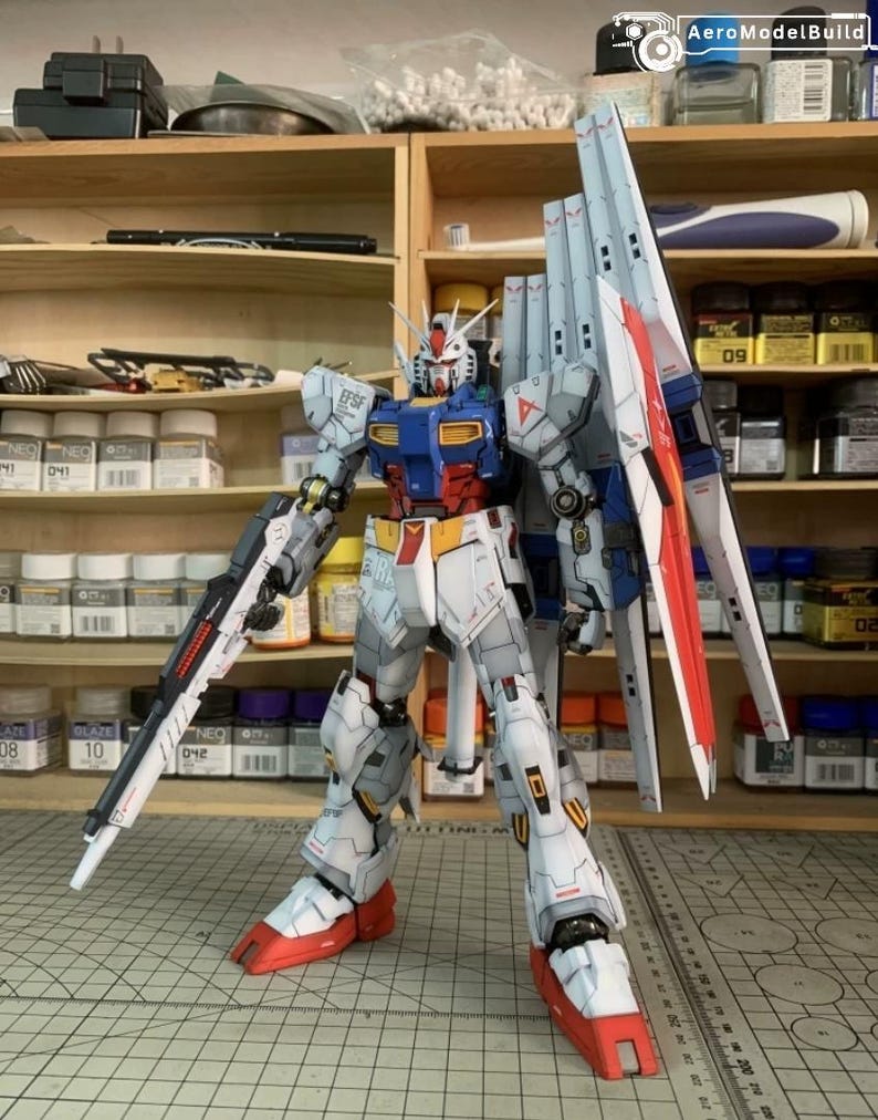 K&ouml;nnte beinhalten: Ein detailliertes Modell eines Gundam-Roboters, haupts&auml;chlich in Wei&szlig;, Blau, Rot und Gelb. Der Roboter ist mit einer gro&szlig;en Waffe und Fl&uuml;geln posiert und weist komplizierte Linien und Abziehbilder auf. Das Modell steht vor einem Hintergrund von Regalen mit verschiedenen Hobbyartikeln.