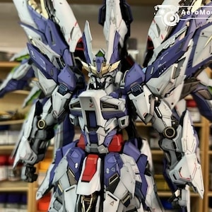 May include: A detailed model robot, predominantly white, purple, and red. It features large wings and intricate mechanical details. The model is set against a blurred background of shelves with paint bottles.