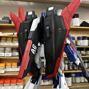 May include: A detailed model kit of a mecha robot, featuring a combination of black, red, white, and blue panels. The model has articulated wings and intricate detailing, with the text "AG ALUCIORE" visible on one of the panels. The model is partially assembled.