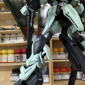 Puede incluir: Maqueta detallada de un robot mecha, con extremidades y paneles articulados. La combinaci&oacute;n de colores incluye tonos verde menta, negro y gris. El modelo est&aacute; montado sobre un soporte, mostrando su pose din&aacute;mica.