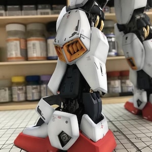 Op de afbeelding: Close-up van een gedetailleerde, gedeeltelijk geassembleerde witte en rode modelrobotpoot. Het model heeft ingewikkelde mechanische details, waaronder oranje en gouden accenten. De voet is felrood en de poot bestaat uit meerdere witte panelen.