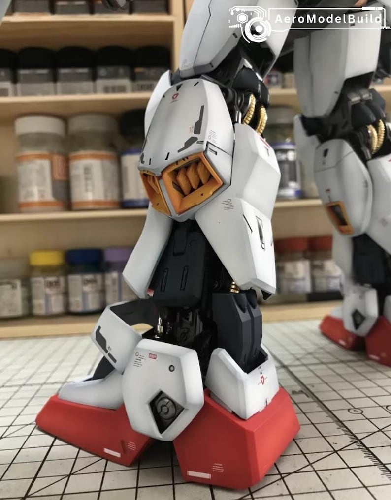 Op de afbeelding: Close-up van een gedetailleerde, gedeeltelijk geassembleerde witte en rode modelrobotpoot. Het model heeft ingewikkelde mechanische details, waaronder oranje en gouden accenten. De voet is felrood en de poot bestaat uit meerdere witte panelen.