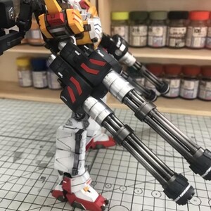 K&ouml;nnte beinhalten: Detailliertes Modell eines Gundam-Roboters mit einer Farbgebung aus Wei&szlig;, Rot, Gelb und Schwarz. Der Roboter ist mit gro&szlig;en, mehrl&auml;ufigen Kanonen ausgestattet und steht auf einer Rasteroberfl&auml;che, die komplizierte mechanische Details zeigt.
