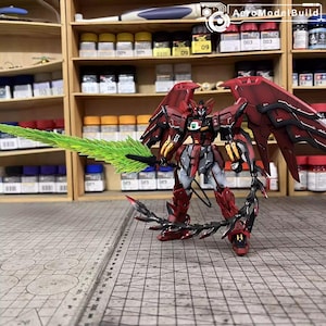 May include: A detailed model of a red and black mecha robot with large wings and a green energy sword. The robot has intricate detailing and is posed on a grid surface. The background includes shelves with paint supplies and the text "AeroModelBuild".