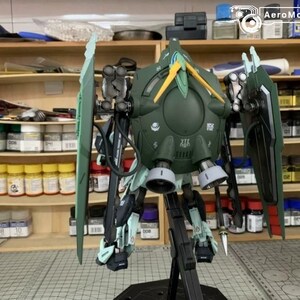 Puede incluir: Maqueta detallada de un robot mec&aacute;nico verde y negro con detalles en amarillo y verde claro. El modelo tiene grandes estructuras en forma de ala y est&aacute; montado sobre un soporte negro. El fondo muestra estantes con suministros de pintura.