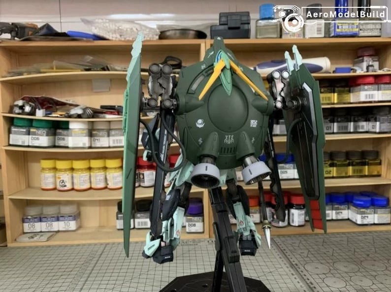 Puede incluir: Maqueta detallada de un robot mec&aacute;nico verde y negro con detalles en amarillo y verde claro. El modelo tiene grandes estructuras en forma de ala y est&aacute; montado sobre un soporte negro. El fondo muestra estantes con suministros de pintura.