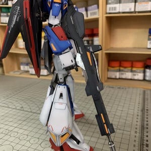 May include: A detailed model robot with a gun, predominantly white, blue, red, and black, with orange detailing. It features large wings and a long gun. The model is on a cutting mat with metric measurements.