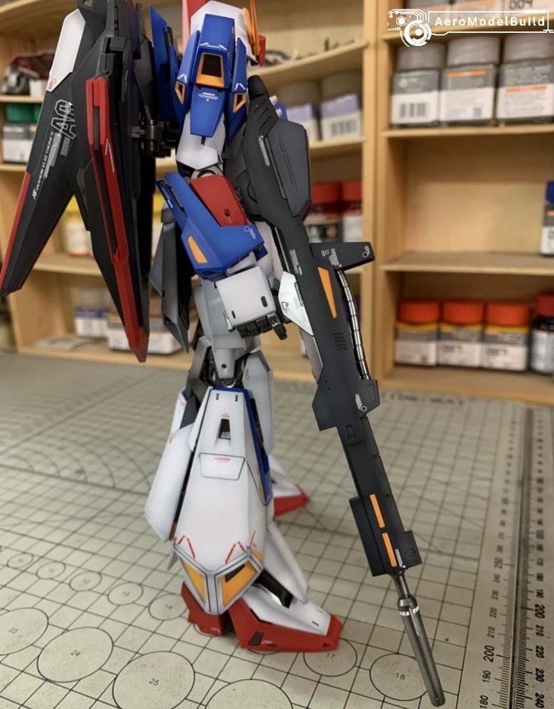 May include: A detailed model robot with a gun, predominantly white, blue, red, and black, with orange detailing. It features large wings and a long gun. The model is on a cutting mat with metric measurements.