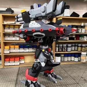 May include: A detailed model of a futuristic robot with a complex design. The model features a combination of gray, black, and red components, with multiple missile launchers and jet-like wings. The model is on a cutting mat.