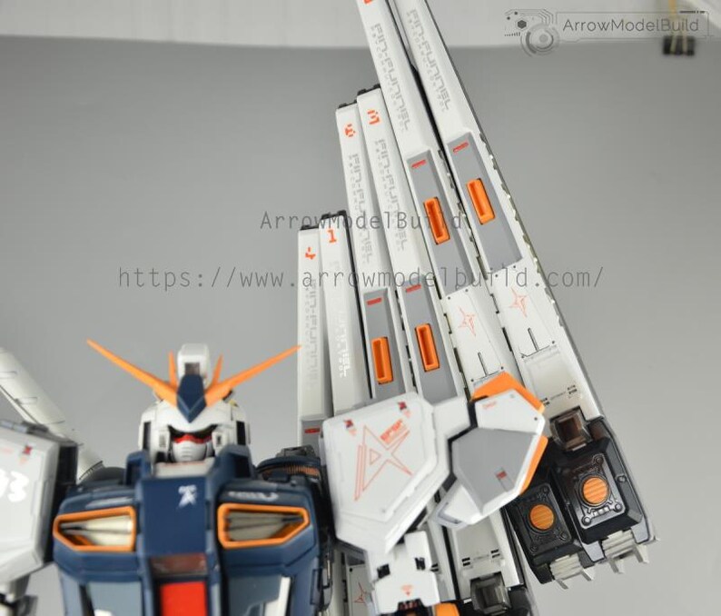 May include: A detailed model kit of a Gundam robot, featuring a white, navy blue, and orange color scheme. The model has intricate paneling and markings, with text visible on the white panels. The robot's head has orange antennae.