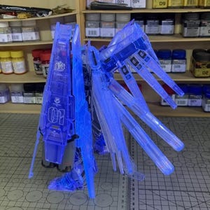 May include: A transparent blue model kit, possibly a Gundam, with intricate wing-like structures and a central body. The model features detailed panel lines and is illuminated, showcasing its internal components. The number "01" is visible on the model.