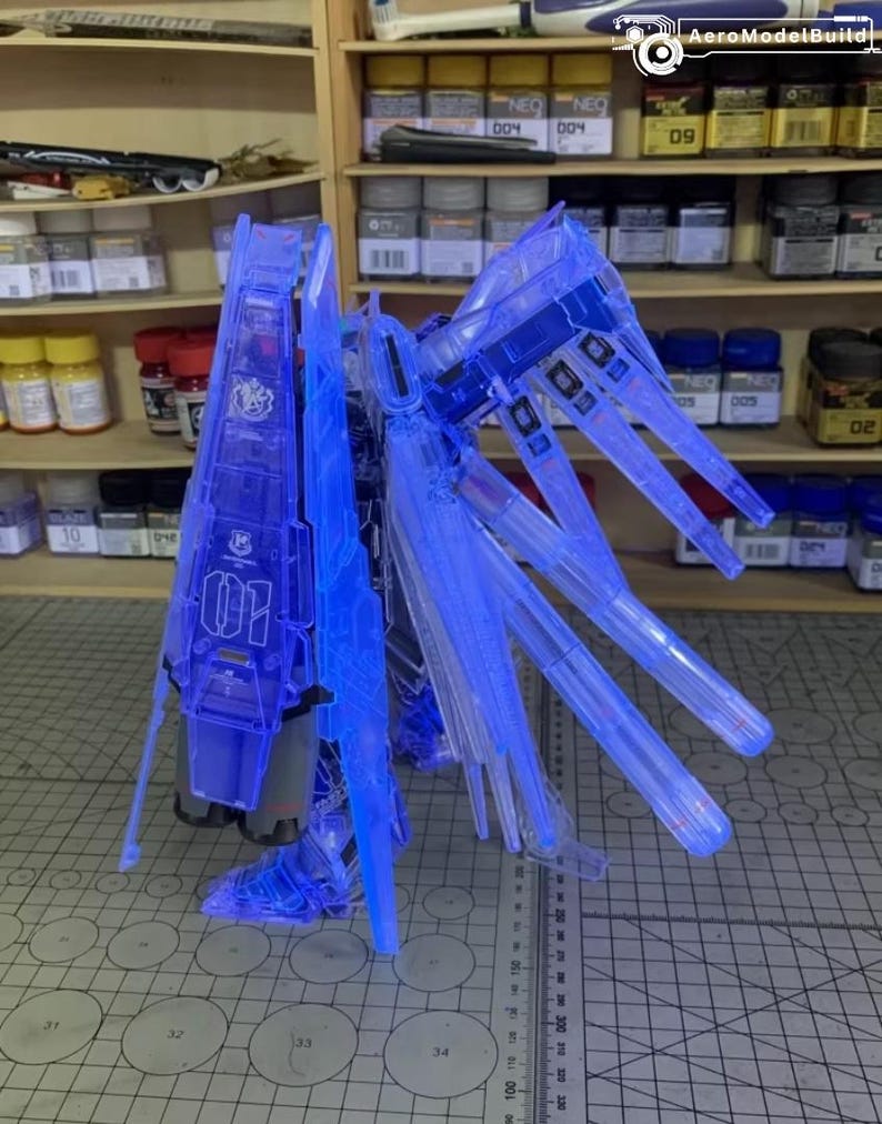 May include: A transparent blue model kit, possibly a Gundam, with intricate wing-like structures and a central body. The model features detailed panel lines and is illuminated, showcasing its internal components. The number "01" is visible on the model.