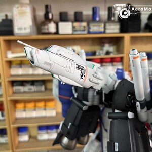 May include: A partially assembled model robot kit, predominantly white, blue, and black. The model has detailed features, including cylindrical boosters and text. The background shows shelves with paint bottles and other modelling supplies.