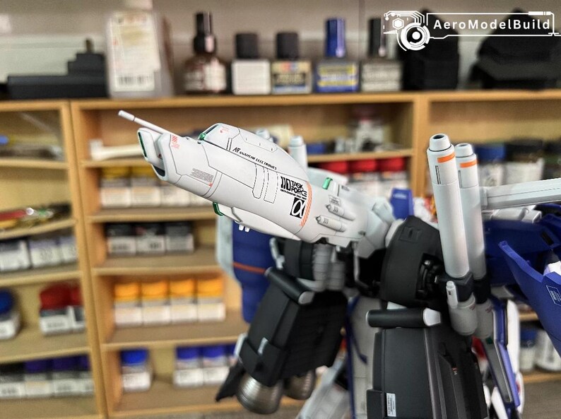 May include: A partially assembled model robot kit, predominantly white, blue, and black. The model has detailed features, including cylindrical boosters and text. The background shows shelves with paint bottles and other modelling supplies.