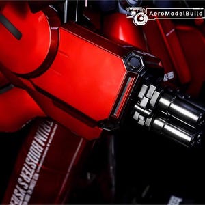 May include: Close-up of a red and black model kit component. The image features a metallic red arm with a silver gun barrel and the text "TRIM BOOSTER SYSTEM". The AeroModelBuild logo is visible in the top right corner.