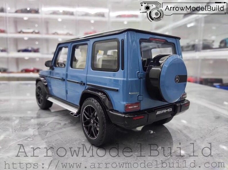 May include: A blue diecast model of a Mercedes-Benz G-Class SUV. The model features black wheels, a spare tire, and AMG badging. The image includes the text "ArrowModelBuild" and a website address.