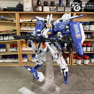May include: A detailed model robot, predominantly white and blue, holding a large gun. The model has intricate mechanical details and is displayed on a clear stand. Shelves with paints and tools are visible in the background.