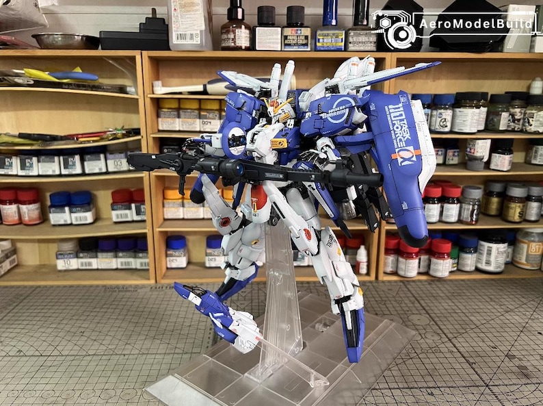 May include: A detailed model robot, predominantly white and blue, holding a large gun. The model has intricate mechanical details and is displayed on a clear stand. Shelves with paints and tools are visible in the background.