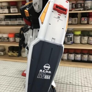 Op de afbeelding: Een gedeeltelijk geassembleerde modelrobot in wit, zwart, rood en geel. Het armschild van de robot toont de tekst "A.E.U.G. AG004" in het wit. Het model staat in een werkplaats met verfpotten op planken op de achtergrond.