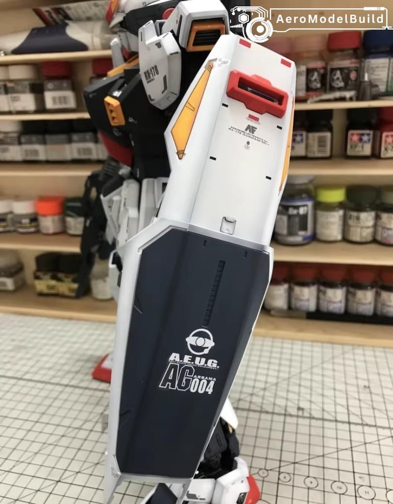 Op de afbeelding: Een gedeeltelijk geassembleerde modelrobot in wit, zwart, rood en geel. Het armschild van de robot toont de tekst "A.E.U.G. AG004" in het wit. Het model staat in een werkplaats met verfpotten op planken op de achtergrond.